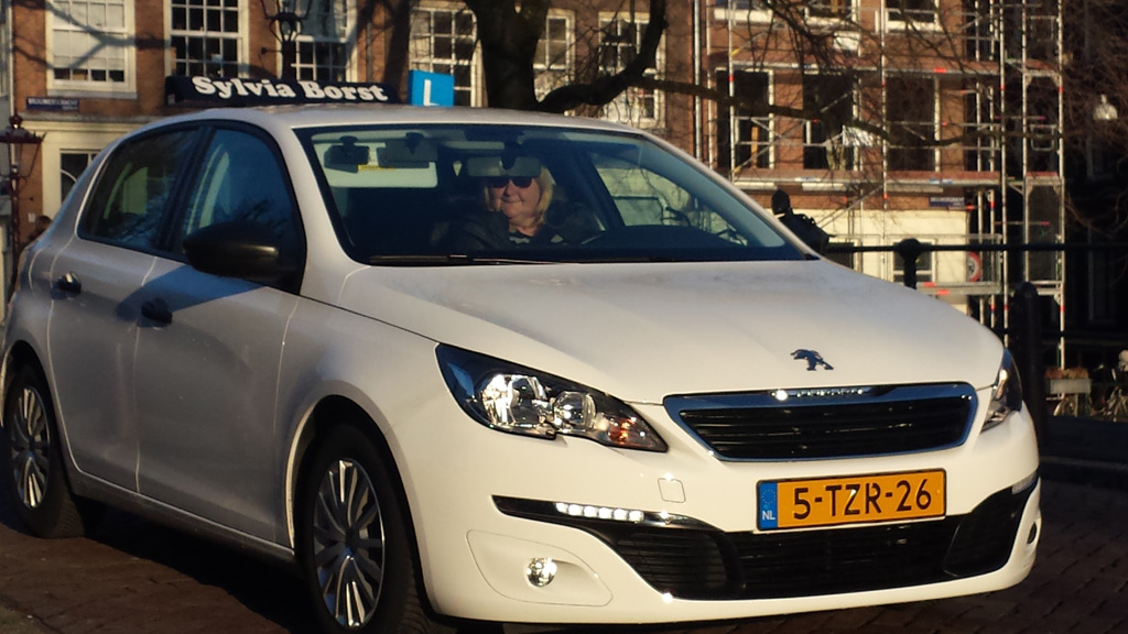 Sylvia Borst in haar witte lesauto met 'Sylvia Borst' bovenop, voor een gracht in Amsterdam